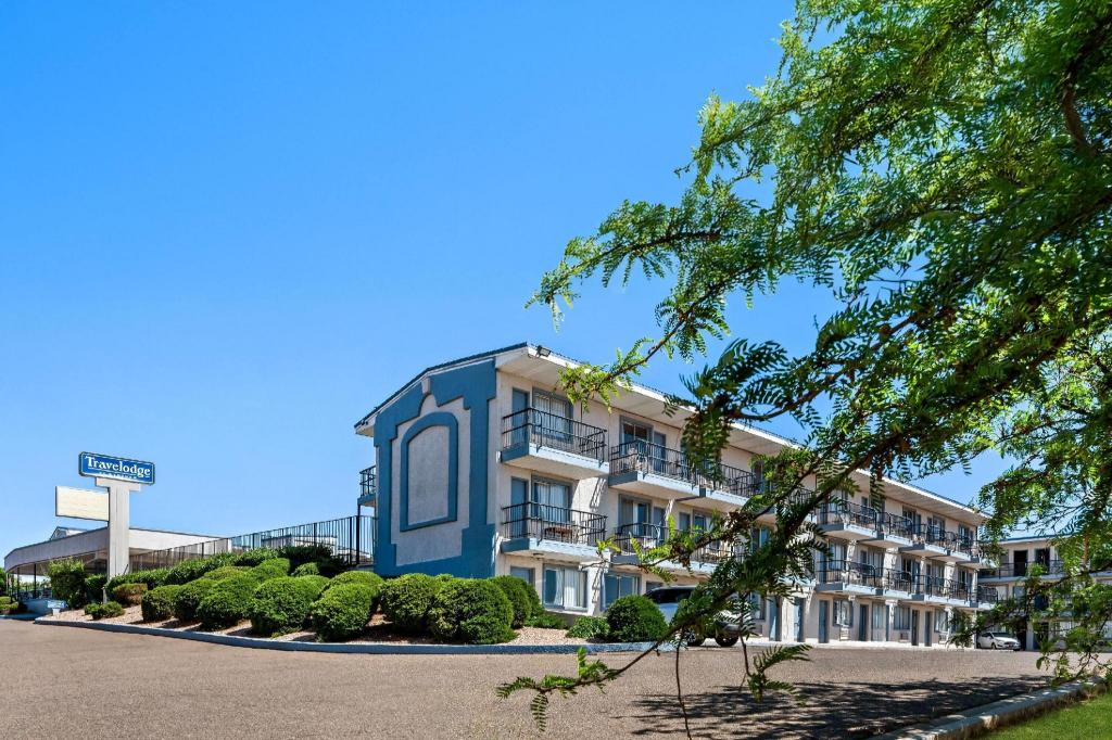 Three-story motel building with balconies, blue and white exterior, surrounded by trimmed bushes, under a clear blue sky. A Travelodge sign is visible in the background.
