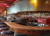 A curved wooden bar with hanging glassware, bar stools, bottles, and a register in a restaurant or lounge with red walls and ambient lighting.