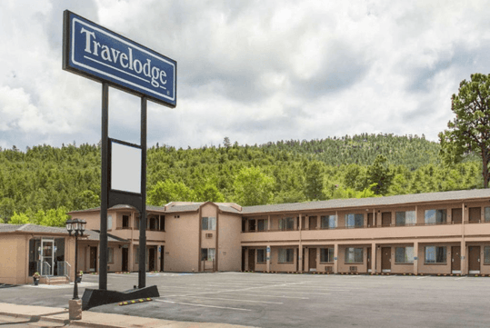 Travelodge motel with beige buildings, surrounded by trees and hills. Large sign in parking lot. Cloudy sky overhead.