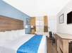 Hotel room with a king-sized bed, blue accent wall, wooden headboard, desk with chair, wall-mounted TV, window with curtains, and framed art on walls.