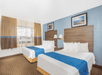 Hotel room with two double beds featuring blue and white bedding, a window with curtains, framed art above headboards, wall sconces, and beige carpet.