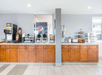 Breakfast area with wooden cabinets, coffee and juice dispensers, waffle maker, cereals, muffins, and pastries displayed on counters against a light gray wall.