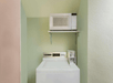 A washing machine is placed below a microwave oven on a wall-mounted shelf, set against pastel-colored walls.