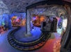 A dimly lit indoor amusement ride with tracks winding through a cave-like setting. The ride has mining-themed decorations and colorful lighting effects.