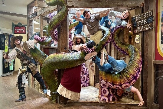 A group of people dressed in costumes are playfully posing with large tentacles in a themed setting, including a sign that says "Don't feed the Kraken.