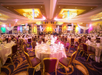 A decorated banquet hall with round tables set for an event. Tables feature white tablecloths, glassware, and folded napkins. Warm lighting and drapery enhance the elegant atmosphere.