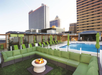 A rooftop terrace with green seating around a fire pit, overlooking a pool. Tall buildings are visible in the background.