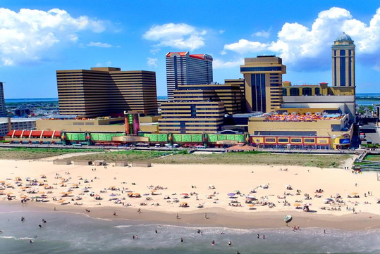 Aerial view of a beach with people sunbathing, adjacent to a cityscape featuring tall, colorful buildings under a partly cloudy sky.