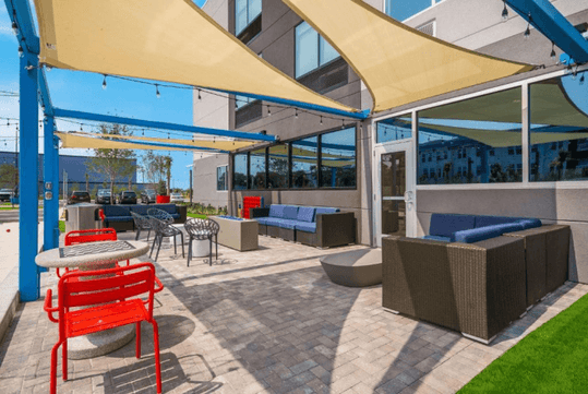 Outdoor patio with red and gray seating, shaded by sail canopies. Pavers cover the ground, and a building is nearby.