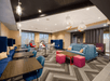 A modern lounge with wooden tables, blue sofas, and pink ottomans. Overhead lighting and large windows create a bright atmosphere. Geometric carpet design enhances the contemporary feel.