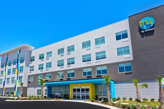 Four-story Tru by Hilton hotel with a bright facade, featuring a blue and yellow entrance and palm trees in front, set against a clear blue sky.