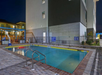 Outdoor swimming pool at night, surrounded by a fence with a building in the background. The pool area is empty, and there are signs on the fence.