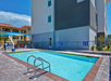 Outdoor pool next to a building, surrounded by a fence. The area includes a ladder for entry and exit, safety signage, and loungers visible in the background.