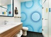 Modern bathroom with blue dotted wallpaper, a glass shower, wood vanity, and wall mirror. A brown bag hangs on the wall, and towels are neatly stacked on a shelf.