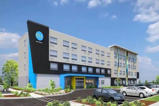 A modern hotel with a blue and black facade, featuring four stories and a parking lot with several cars in front.