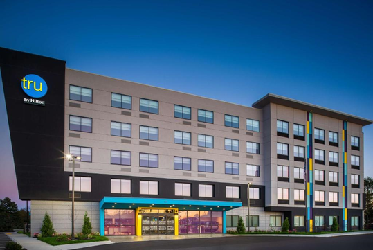 Five-story hotel building with a modern facade, featuring large windows and a prominent entrance. The sky is clear during early evening.