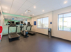 Fitness room with treadmills, ellipticals, and wall-mounted mirrors. Large windows allow natural light. Walls are light-colored with ceiling lights and emergency equipment visible.