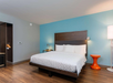 Modern hotel room with a large bed, wooden floor, blue accent wall, bedside table, orange round side table, and standing lamp.