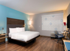 A modern hotel room with a large bed, wooden floor, desk, chair, and a window with a roller blind featuring a subtle pattern. Walls are painted in light blue and gray tones.