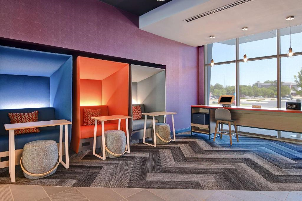 Modern seating area with three colorful, semi-enclosed booths, small tables, and cushioned stools. Large windows on the right let in natural light, illuminating the patterned carpet.