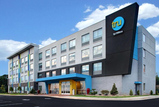 Four-story hotel with a modern facade, featuring a mix of gray and blue colors, marked by the "Tru by Hilton" logo on a corner. Parking lot and landscaped greenery in the foreground.