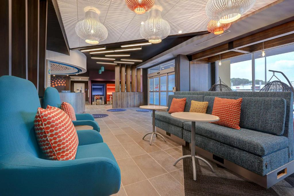 Modern hotel lobby with blue and gray seating, round tables, patterned pillows, and pendant lights. Geometric ceiling design and large windows on the right.