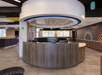 Hotel reception area with a circular wooden front desk, overhead lighting fixture, and patterned wall design. Large windows provide natural light.