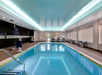 Indoor swimming pool with a chair lift, surrounded by lounge chairs and windows. The ceiling has recessed lighting, and safety signage is visible on the wall.
