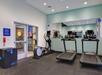 Small gym with two treadmills, an elliptical machine, a workout box, and mirrored wall. A windowed door leads to an adjacent area. Ceiling lights illuminate the space.