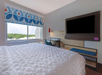 Hotel room with a bed, desk, chair, large TV, and window with a patterned curtain. The view outside shows trees and buildings.