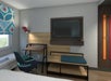 A hotel room with a desk and chair, wall-mounted TV, and shelves. A window with a patterned curtain is on the left, and a bed with white linens is partially visible in the foreground.