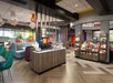 Modern convenience store interior with a self-serve coffee station, seating area, and shelves stocked with various products. Bright lighting, wood flooring, and large windows are visible.
