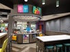Modern convenience store interior with a circular counter displaying snacks and drinks, digital screens above, colorful stools, and geometric patterns on the walls.