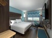 Hotel room with a large bed, wall-mounted TV, desk, chair, and a window with patterned curtains. The walls are painted in blue tones.