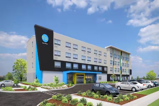 A modern four-story hotel with a blue and white facade, surrounded by parked cars and landscaping, under a partly cloudy sky.