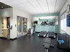 Modern gym with treadmills, stationary bike, weight bench, and exercise equipment. Mirrors on the wall reflect machines. Wall clock and poster visible. Brightly lit interior.