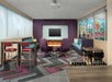A lounge area with blue and purple couches and chairs, brown tables and chairs, windows on the left and right and a purple accent wall with a TV on the back.