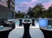 A patio with black standing tables  with candles on them and a buffet to the left and a swimming pool in the background at dusk.