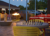 Outdoor patio at dusk with a chessboard on a table, yellow and red chairs, a fire pit, and a large Jenga game nearby. Cars are parked in the background.