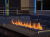 A modern outdoor fire pit with tall flames, enclosed by glass panels, surrounded by a dark seating area and colorful wall background.
