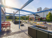 Outdoor patio with red chairs, a fire pit, and string lights under a blue pergola. Hotel building and entrance are visible in the background.