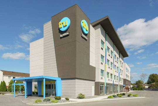 Four-story modern hotel with gray and white facade, blue accents, and "tru" logo. Entrance has a blue awning. Clear sky and trees in the background.