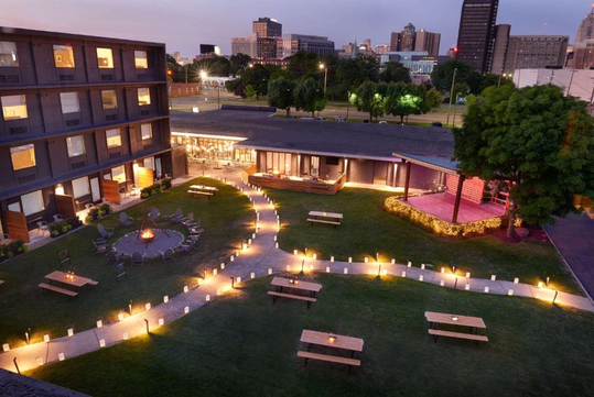 A courtyard with lit pathways, picnic tables, a fire pit, and a stage is surrounded by modern buildings against a city skyline at dusk.