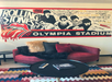 A red couch with cushions is positioned in front of a wall mural featuring vintage "Rolling Stones 1964 Tour" artwork at Olympia Stadium. A round coffee table is in the foreground.