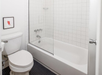 A bathroom featuring a white toilet, a bathtub with a glass door, white tiled walls, and a small piece of artwork on the wall.