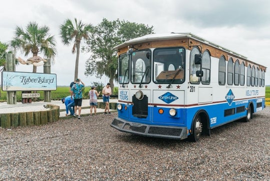 A blue and white trolley labeled "Kelly Tours" is parked near a sign that reads "Tybee Island." People stand nearby on a gravel path with palm trees in the background.