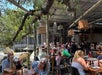 Outdoor restaurant with people dining under a canopy of trees and hanging decorations. Tables are filled with patrons enjoying meals in a lively atmosphere.