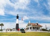 A white and black lighthouse stands next to a historic house with a picket fence under a blue sky. An American flag flies nearby.