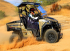 A person drives a camouflage-patterned off-road vehicle through a dusty desert landscape.