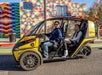 A man driving a GoCar with a woman in the seat behind him parked on the side of a road in front of a colorful building.